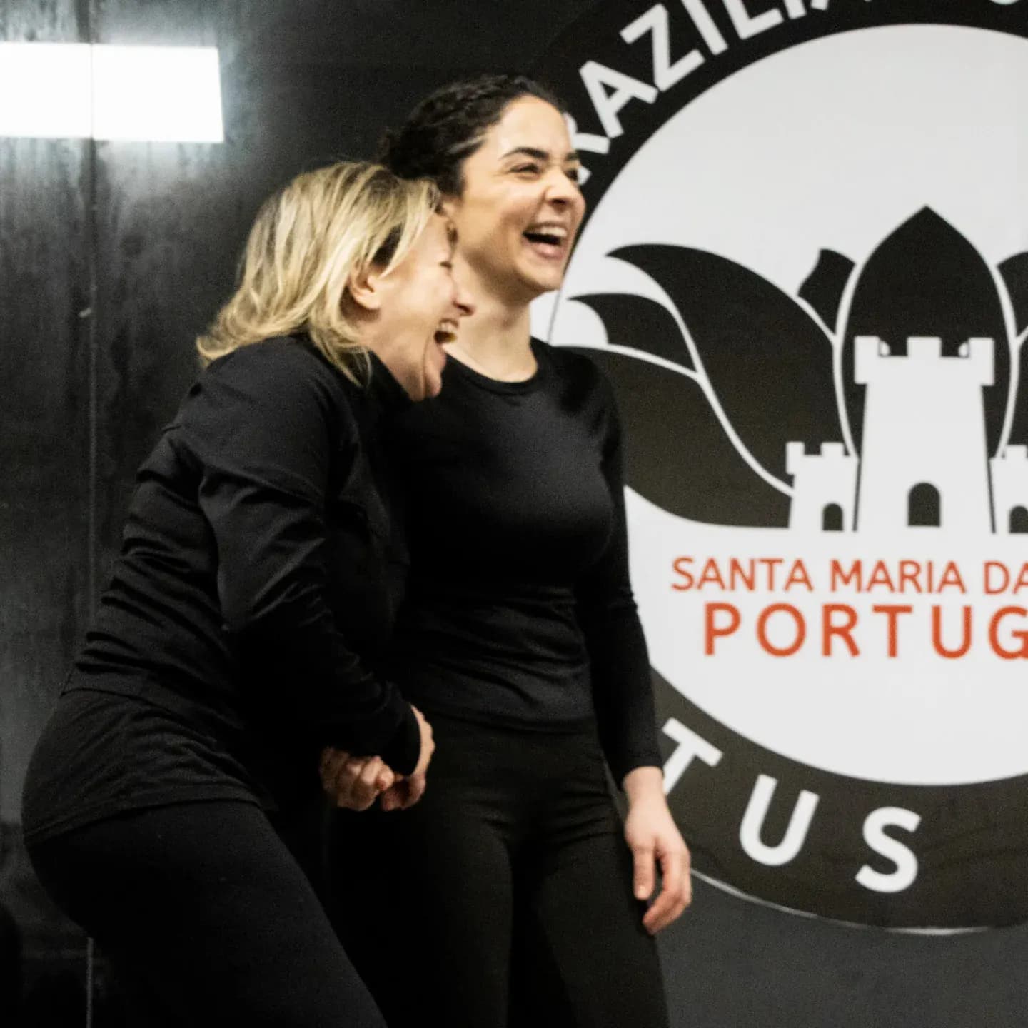 Duas alunas a rir durante o seminário de defesa pessoal para mulheres, com o logótipo da academia atrás
