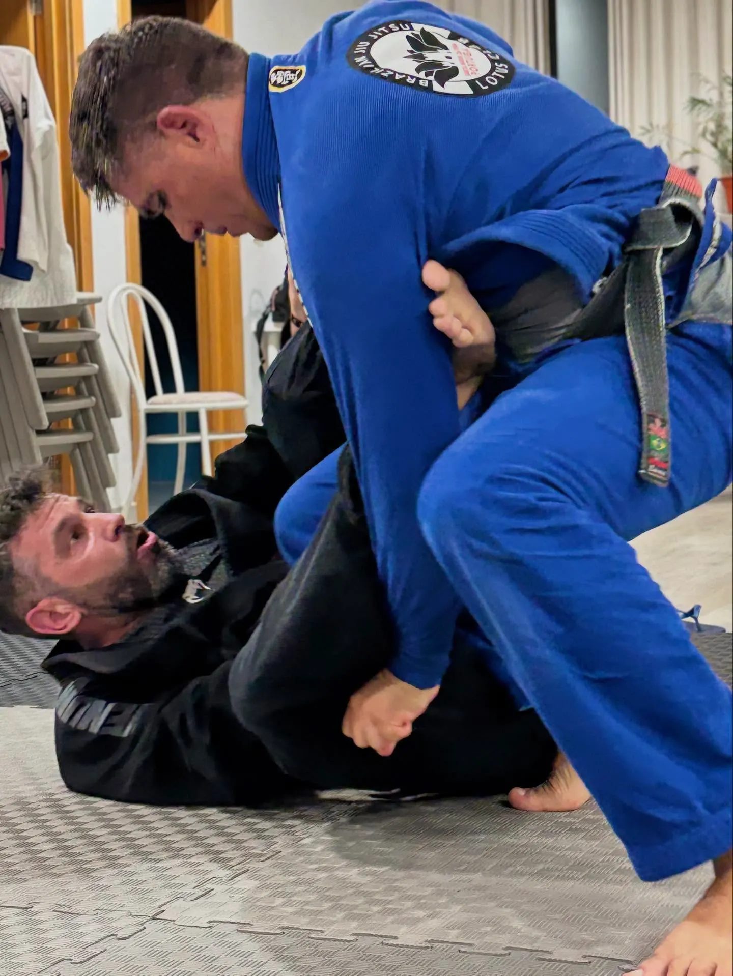 Treino de jiu-jitsu no tatame, um aluno de azul sobre a guarda de outro de preto